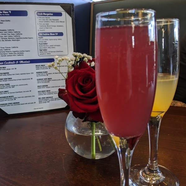 Two cocktails, one red and one yellow, with a red rose and baby's breath on a table.