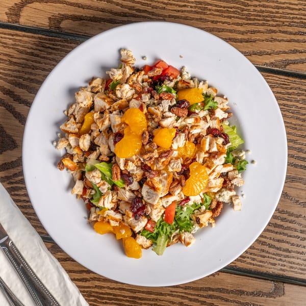 A vibrant salad with grilled chicken, mandarin oranges, pecans, and cranberries on a white plate.