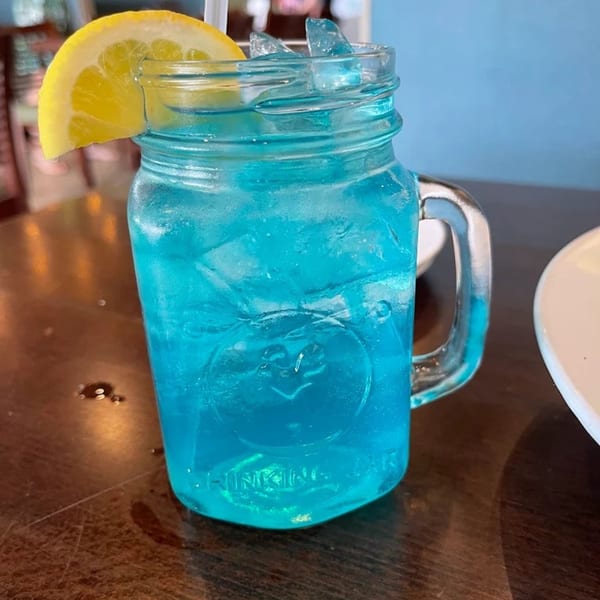 A vibrant blue drink with ice and a lemon slice in a mason jar on a wooden table.